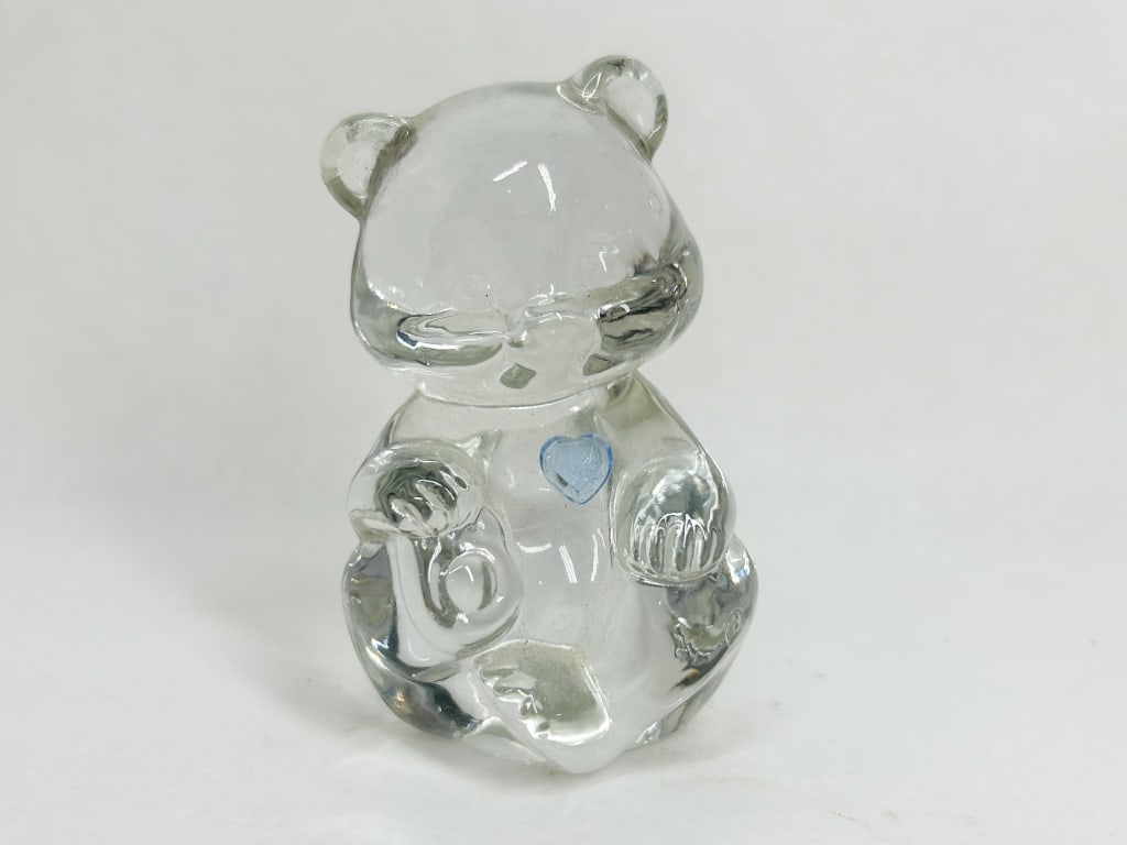 FENTON CLEAR GLASS BIRTHSTONE BEAR: A clear pressed glass figurine of a seated teddy bear featuring a small raised blue heart on the chest. An adhesive silver paper label on the base depicts a stylized script F within an oval above the 