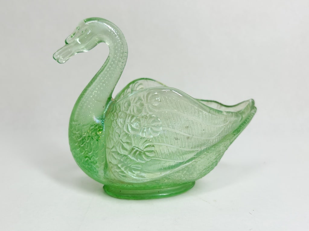 GREEN PRESSED GLASS SWAN DISH: A green pressed glass open salt or nut dish in the form of a swimming swan. The piece features stippled texture on the neck and stylized feathers on the wings, set upon an oval pedestal base. No maker