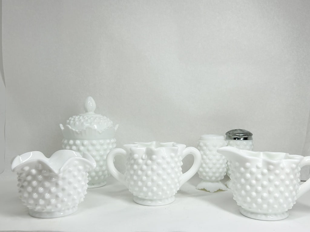 A COLLECTION OF WHITE HOBNAIL MILK GLASS TABLEWARE: A set of six white milk glass items featuring a raised hobnail pattern throughout. The collection includes a lidded sugar bowl with a ruffled rim and finial, a creamer with an applied handle, a double