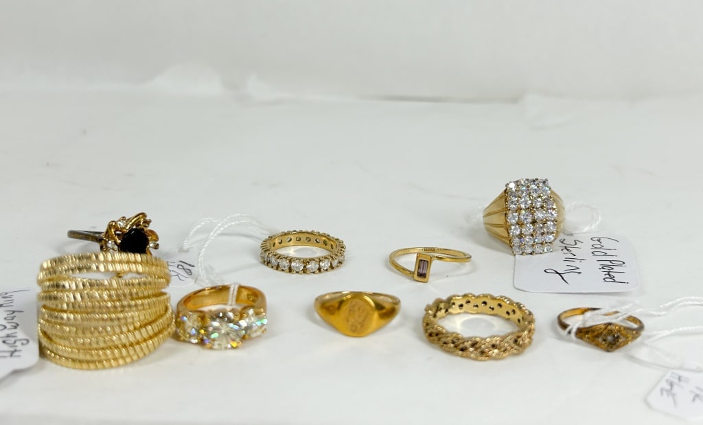 COLLECTION OF NINE GOLD PLATED RINGS: An assortment of nine gold-tone rings including cocktail, signet, and eternity styles. The collection features a ten-row Tubogas "Highway" ring, a three-stone basket-set ring, a black stone cluster ri