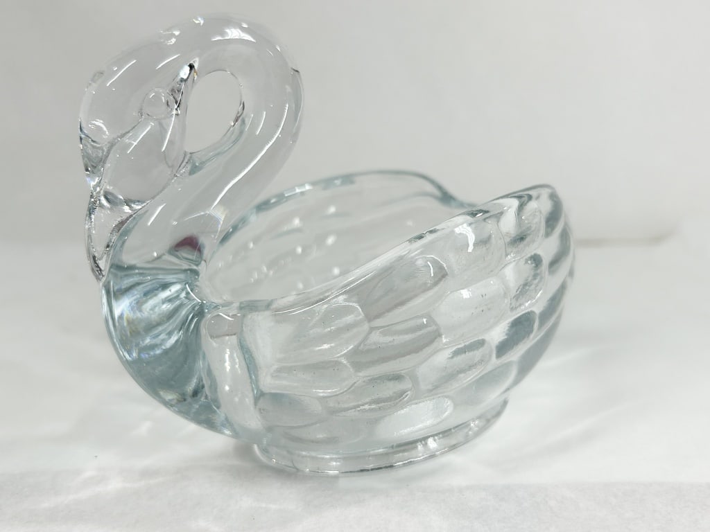 PRESSED GLASS SWAN TRINKET DISH GLOWS: A clear, press-molded glass swan-form dish featuring a feathered wing motif and a recurved neck handle. A hand-written paper label on the interior base reads Glows; the piece exhibits a pale green