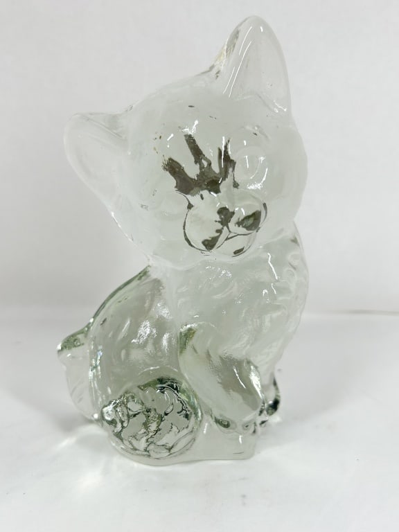CLEAR GLASS SITTING KITTEN FIGURINE: A cast colorless glass figurine of a seated kitten featuring a textured surface and dark applied pigment to the eyes and nose. A small square of white adhesive residue is present on the back of the he