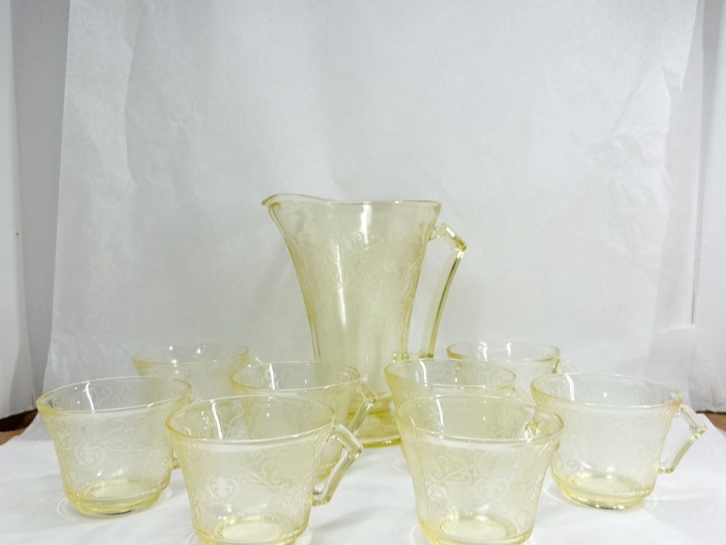 YELLOW DEPRESSION GLASS PITCHER AND CUPS: A set of depression-era pressed glass in a transparent yellow color featuring the Florentine Poppy pattern. The collection includes a footed pitcher with a flared rim and angular handle, accompanied b
