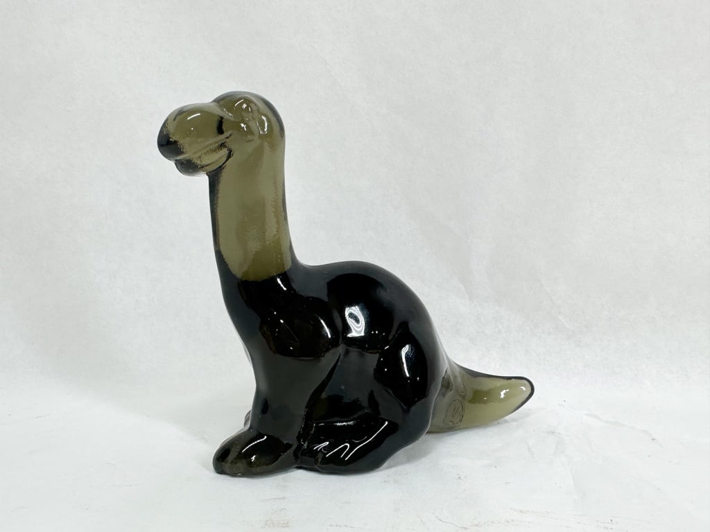 TWO-TONE SMOKY GLASS DINOSAUR FENTON: A stylized glass figurine of a long-necked dinosaur depicted in a seated pose. The piece features a bi-color palette with a dark charcoal body transitioning to translucent smoky-olive glass at the nec