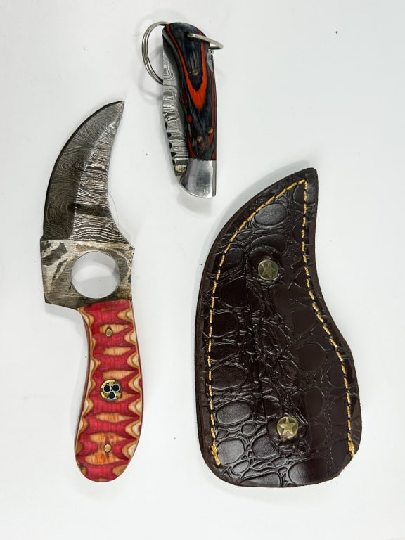 TWO DAMASCUS STEEL KNIVES AND LEATHER SHEATH: Two custom knives including a fixed blade skinner and a lockback folding pocket knife, both featuring pattern-welded Damascus steel blades. The skinner is constructed with a red and orange laminated p