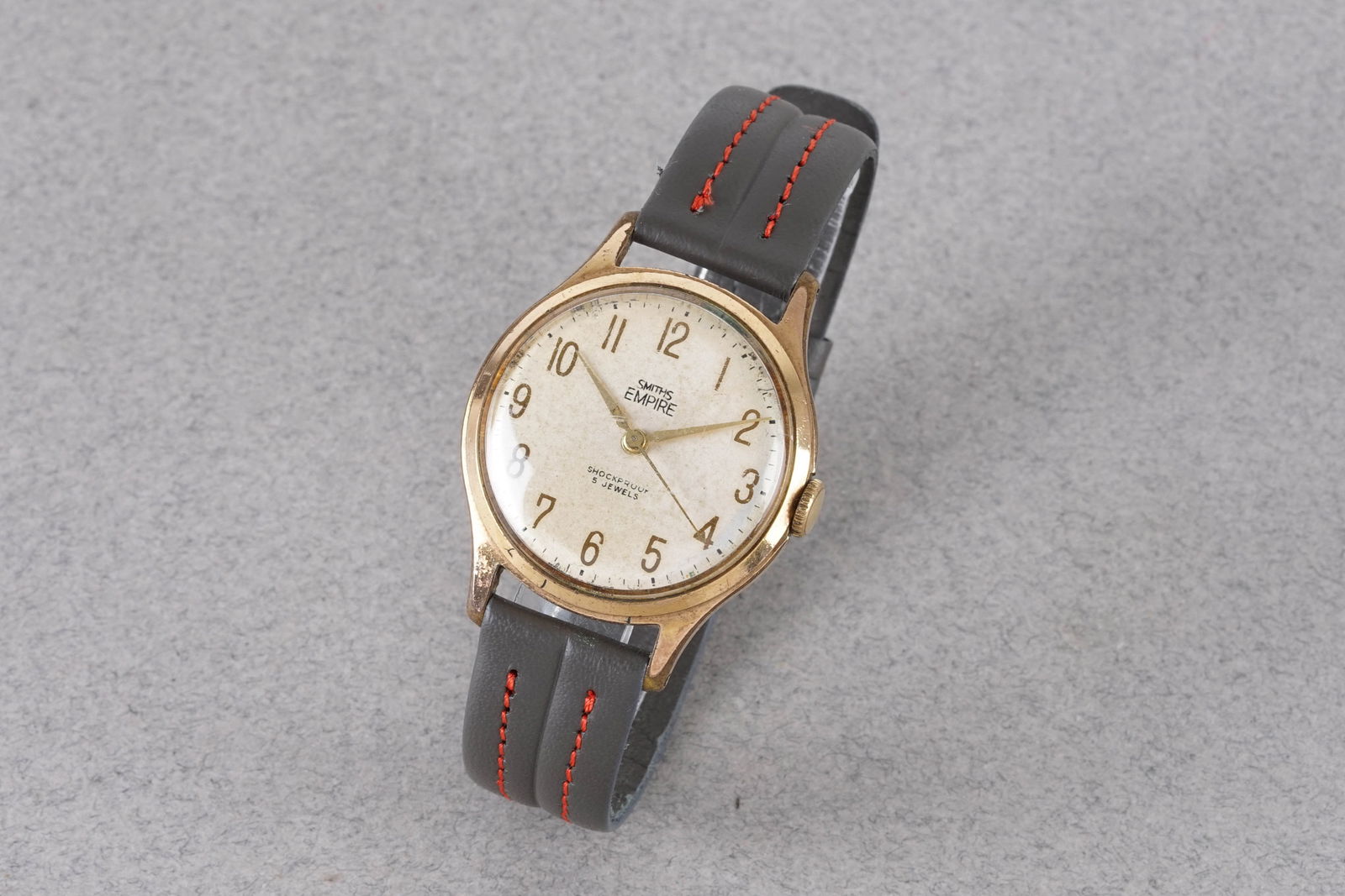 SMITHS EMPIRE GOLD PLATED WRISTWATCH: SMITHS EMPIRE GOLD PLATED WRISTWATCH, this watch is fitted with a circular patina dial with arabic numeral hour markers and hands. The 33mm gold plated case is fitted with a crown and a case back.