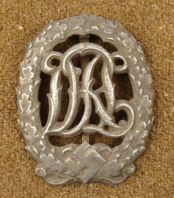 WWII Nazi SA Sports Badge w/ Initials Silver Pin DRGM: This is a WWII era Nazi SA Sports Badge. It is silver with script initials DRL, bordered by an oak leaf pattern. The back is maker marked HENSLER PFORHEIM, D.R.G.M. (followed by numbers). The size is 