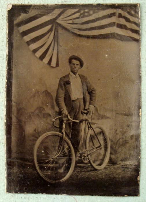 Antique Tin Type Photo Man, Bicycle Bike, American Flag: This antique tintype photograph is a studio shot of a man with a bicycle, with American flags draped above. The bike is a racing type, and the man is wearing a flower on his coat. Its condition is goo
