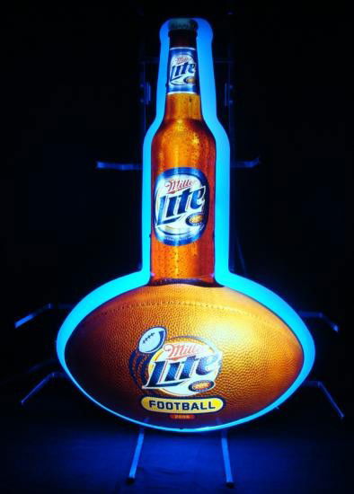 Miller Lite Beer Football Neon Bar Sign Ball Bottle: A big Miller Lite bottle rests on a Miller Lite football, and it's glowing brilliantly. This terrific sign is plastic with realistic litho images, all lit up from behind by light blue neon. It is in e