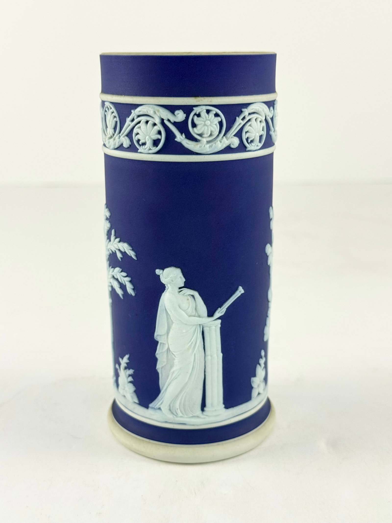 Wedgwood Jasperware Cylinder Vase: A finely executed Wedgwood Jasperware cylinder vase, rendered in the iconic matte cobalt blue ground with applied white neoclassical relief decoration. The tall cylindrical form is adorned with classi