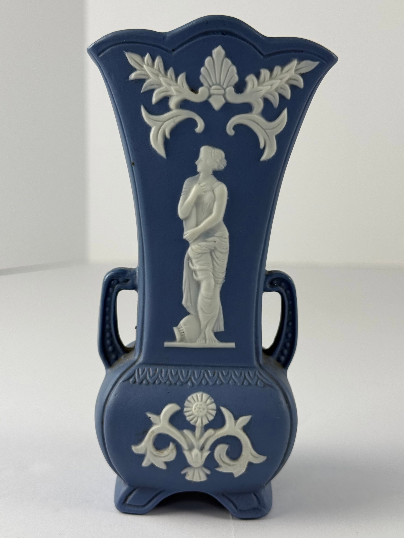 Blue Jasperware / Wedgwood Style Two-Handled Vase: A blue jasperware-style two-handled vase featuring applied white relief decoration of a classical female figure, framed by scrolling foliage and shell motifs. The vase is executed in the traditional m