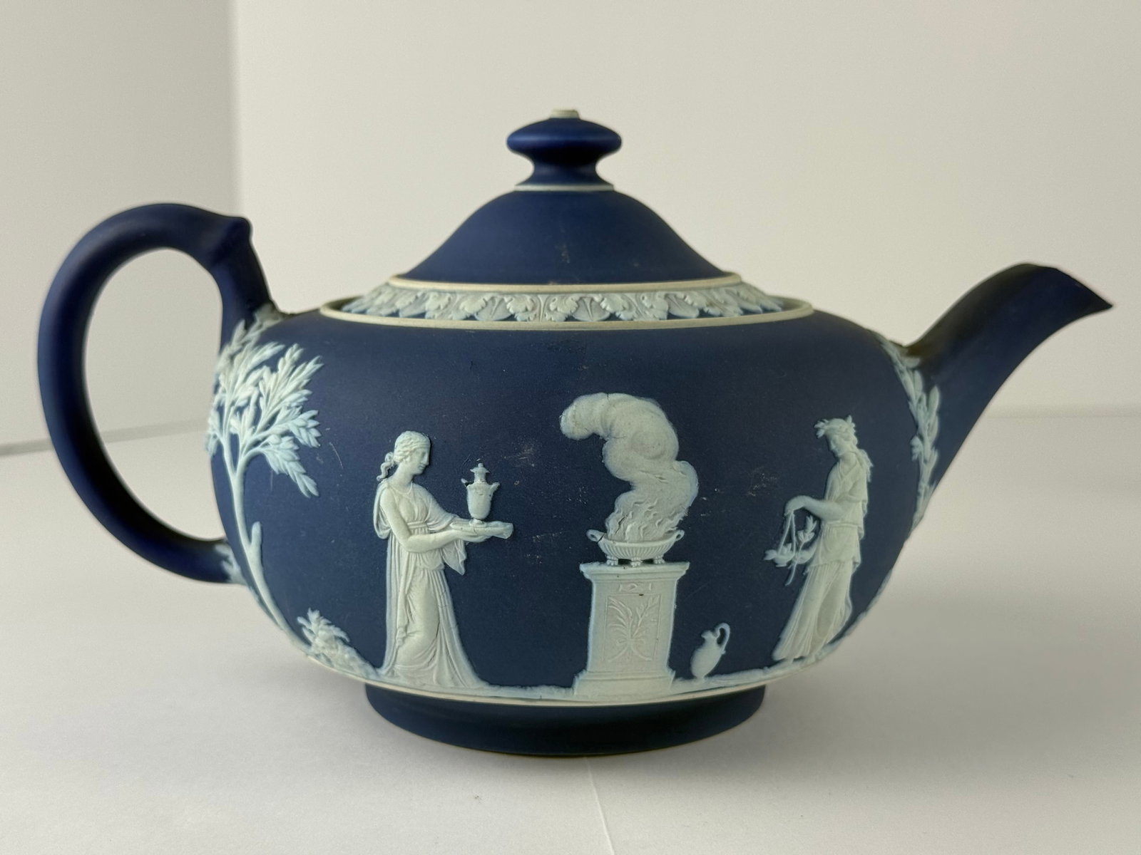 Wedgwood Jasperware Teapot: A finely crafted Wedgwood Jasperware teapot, executed in the iconic deep cobalt blue matte ground with applied white neoclassical relief decoration. The rounded body is encircled with detailed figural