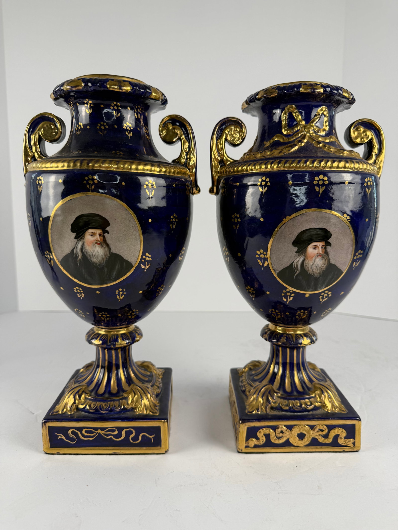 Pair of Cobalt Blue Portrait Urns with Gilt Decoration: An elegant and highly decorative pair of cobalt blue porcelain urns featuring richly gilded accents and finely detailed portrait medallions in the Renaissance taste. Each urn displays hand decorated g