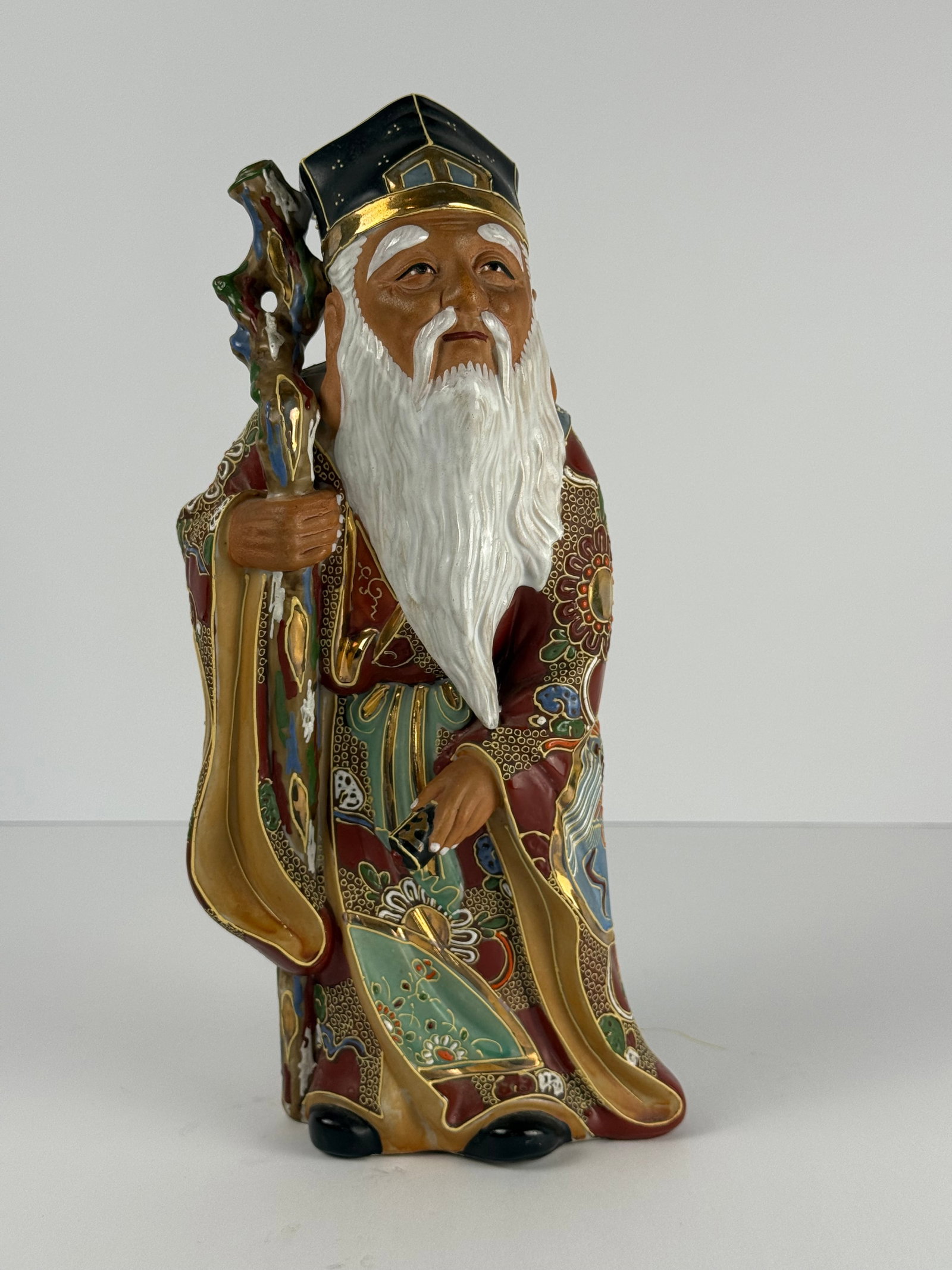 Japanese Moriage Satsuma Style Wise Elder Ceramic Figure: Japanese Satsuma style ceramic figure depicting a wise elder or scholar holding a staff, richly decorated throughout with ornate gilt and enamel floral motifs in the traditional moriage manner. Piece 