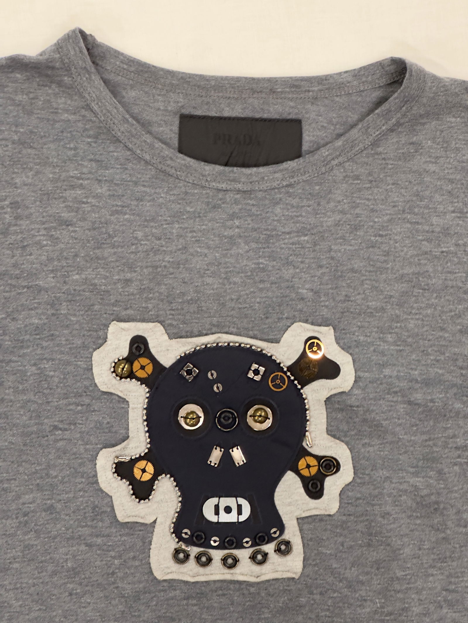 Prada Embellished Gray T-Shirt, Size S: This is a Prada gray T-shirt featuring a unique embellished design on the front. The embellishment includes various metallic and fabric elements, creating a distinctive skull motif. The shirt is craft