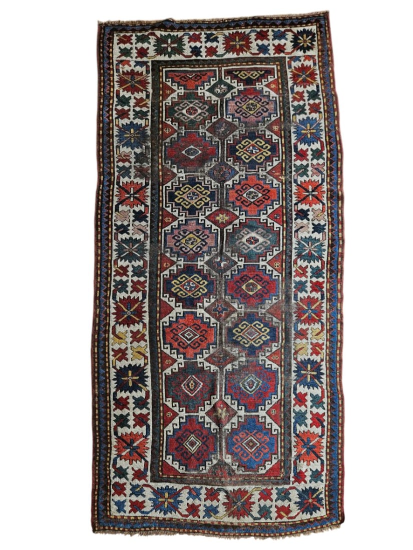 Antique Kazak Rug 4'1" x 9'4": Antique Kazak rug featuring an elongated field with repeating geometric medallions and angular floral motifs, surrounded by multiple patterned borders. Woven in deep red, blue, ivory, rust, and charco