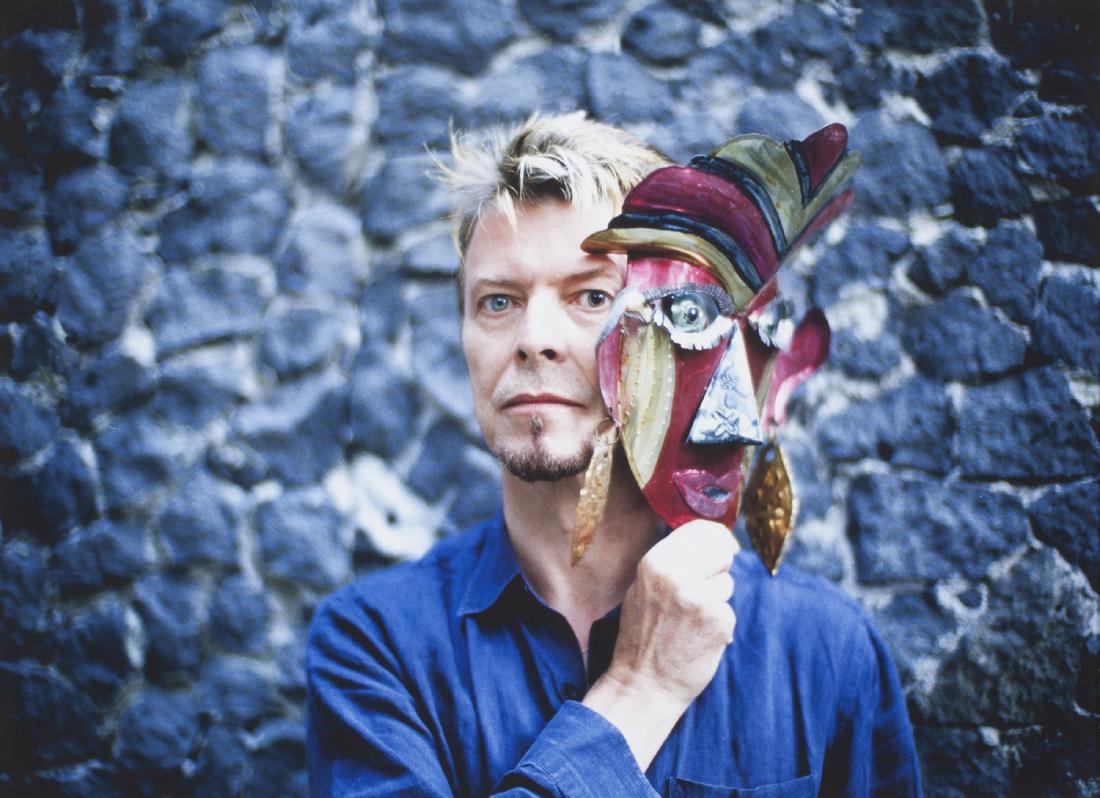 FERNANDO ACEVES (20TH CENTURY): A color photograph of David Bowie holding a mask in front of Frida Kahlo's house in Mexico City, taken in 1997. No visible markings. Not viewed out of the frame. (Glass broken). 14 by 19 inches,