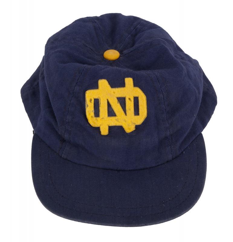 NOTRE DAME CAP: A blue 1940s Notre Dame Fighting Irish cap with a dark green finish under the brim and brown trim inside the cap. On the cap is the logo “ND,” reflecting “Notre Dame.” Football