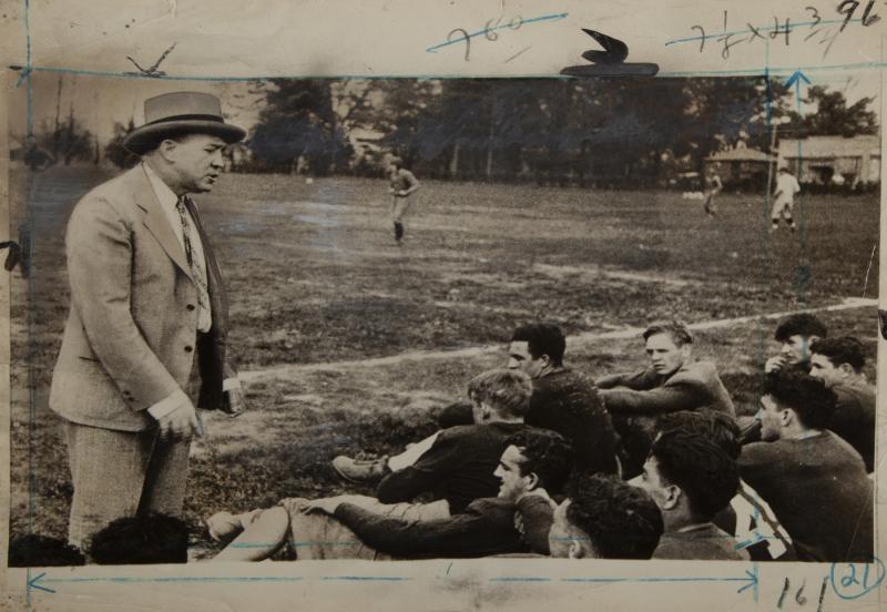 KNUTE ROCKNE PHOTOGRAPH WITH NEWSPAPER CLIPPING: An original black and white photograph of Knute Rockne. In the photograph, Rockne is seen talking to his men during spring training in South Bend, Indiana. Measurement annotations are visible to the f