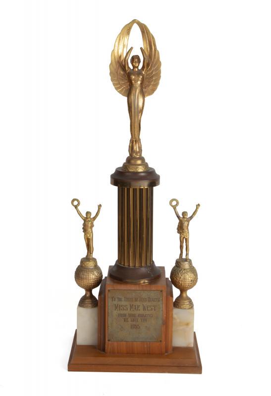 MAE WEST 1955 TROPHY AND PHOTOGRAPH: A gold tone trophy on a wood and marble base with a plaque that reads "To the Queen of Mens Hearts Miss Mae West From your Athletes we love you 1955." Accompanied by an image of Mae West accepting the