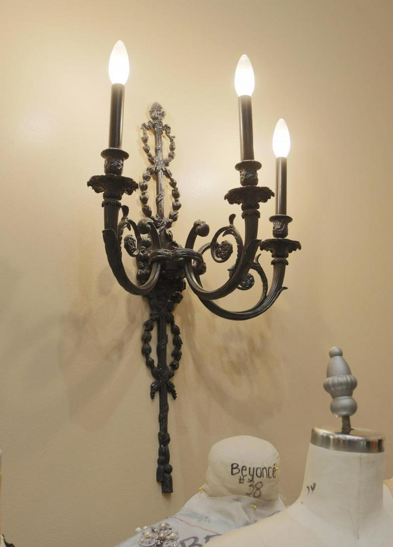 NOLAN MILLER PAIR OF HOLLYWOOD REGENCY WALL SCONCES: A pair of vintage metal black painted three-light sconces with bellflower garlands and tassel terminals. Nolan Miller posed with the wall sconces in the photograph included with the console table. The