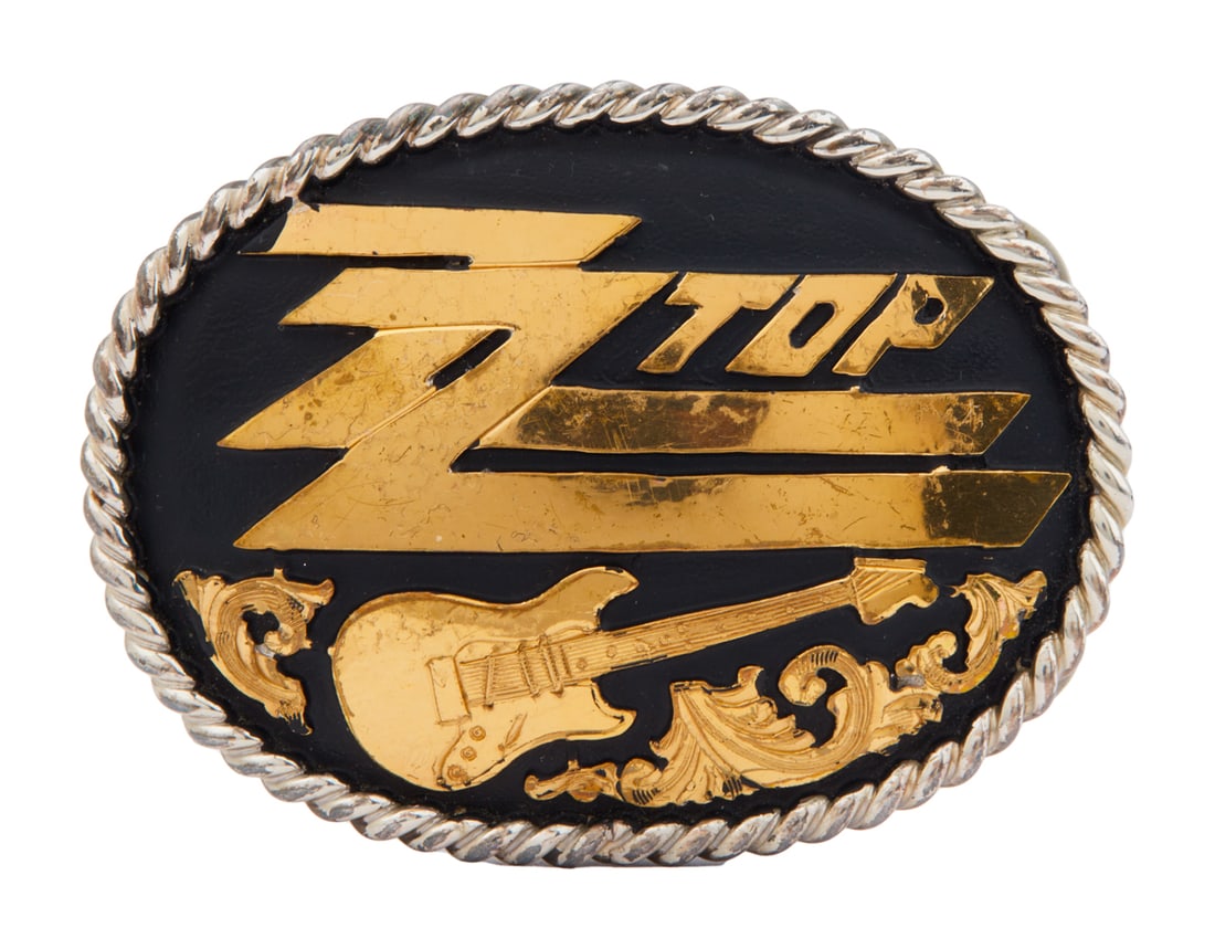 ZZ TOP | DUSTY HILL CUSTOM MONEY CLIP: A brass over blackened metal, oval money clip with "ZZ Top" above a guitar with western scrollwork designs, and rope border. Dimensions: 2 by 1 1/2 inches PROVENANCE From The Estate of Dusty