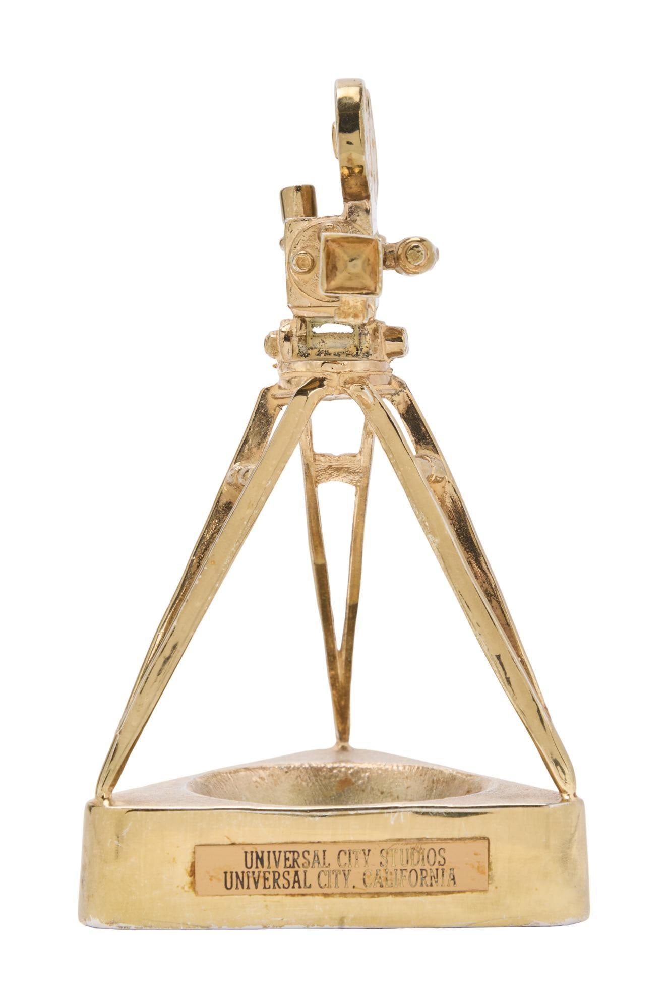 Statuette | Circa 1940s Paperweight from Universal City Studios: A piece made of gold-tone metal; in the form of a movie camera atop a triangular base, plaque on front reads "Universal City Studios / Universal City, California"; likely made for employees