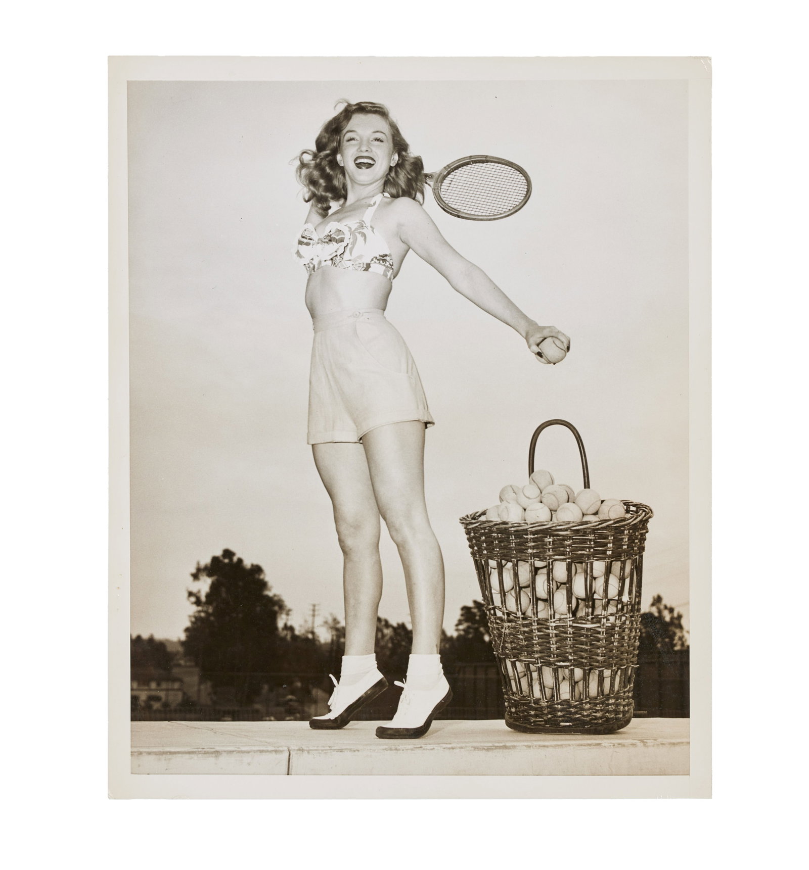 Marilyn Monroe | 1947 Black and White Photograph by Nat Dallinger: An original print; glossy finish, weight-and-a-half paper, depicting the then starlet posing with a tennis racket and a basket of balls; verso with a typed press snipe (once affixed but now loose) has