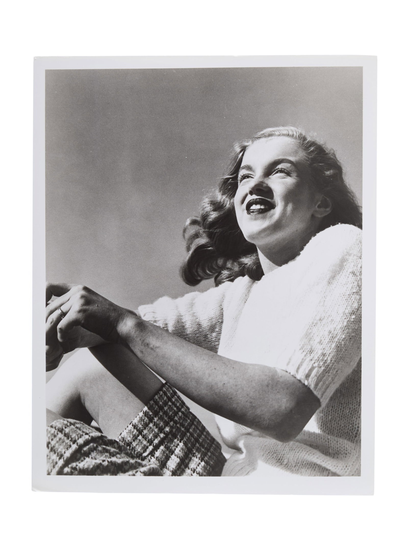 Marilyn Monroe | 1946/1950s/1987 Black and White Photograph by Joseph Jasgur: A circa 1950s later print; glossy finish, depicting the young model smiling as she wears a sweater and plaid shorts; verso with a black ink credit stamp reading "Joseph Jasgur / Photography / Hollywoo