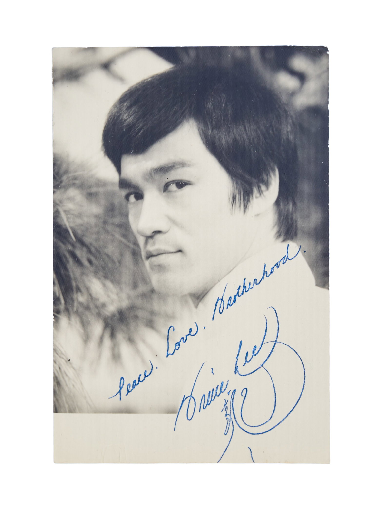 Bruce Lee | Signed Photograph: A small, black and white photograph of and signed by Bruce Lee. The inscription above the signature in blue ink reads, "Peace. Love. Brotherhood." 