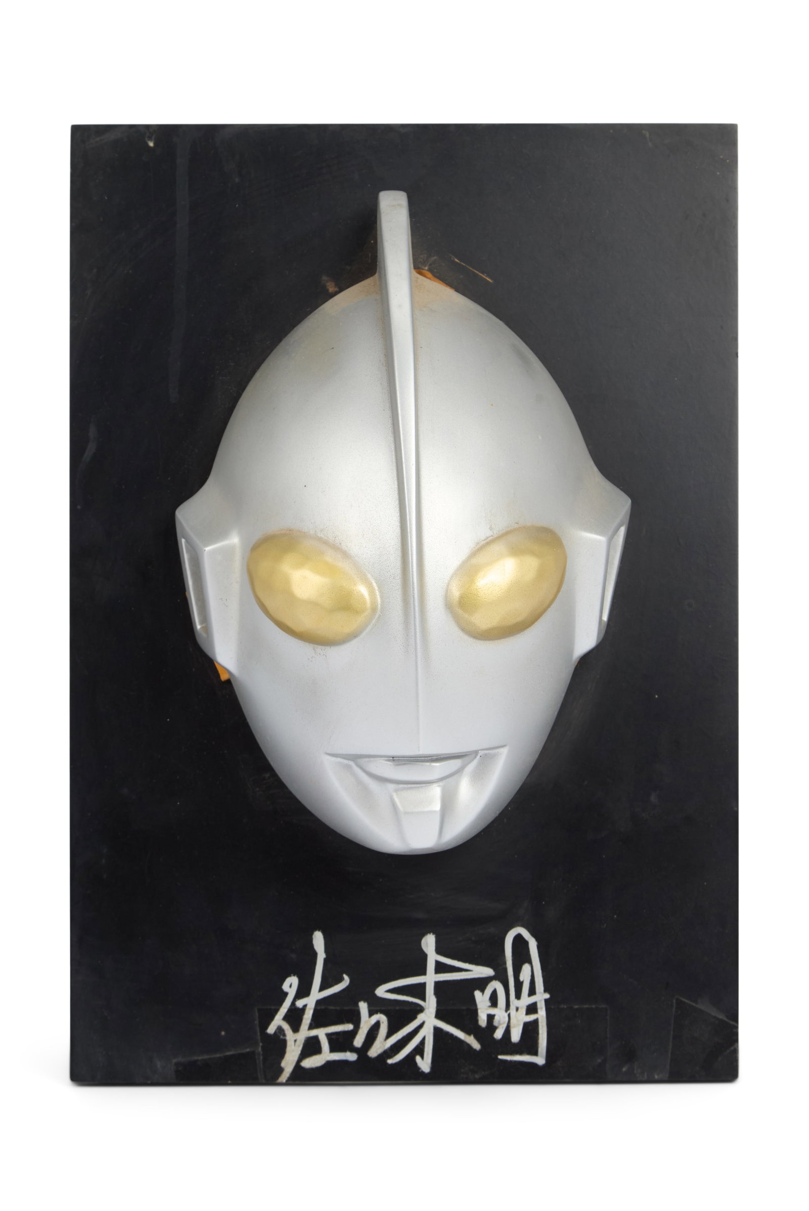 Ultraman | 3-D Mask Display with Akira Sasaki Attributed Signature: A three-dimensional display featuring an Ultraman mask affixed to a black board. A signature in silver marker is on the board beneath the mask. The signature is attributed to original co-designer of