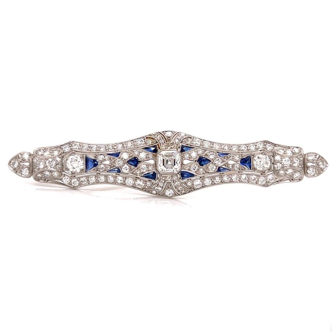 Platinum Art Deco Brooch with Diamonds and Synthetic Sapphires: Platinum Art Deco Brooch with Diamonds and Synthetic Sapphires This Art Deco brooch is crafted in platinum and features a central approximately 1.00 carat Asscher-cut diamond. The brooch is further ad