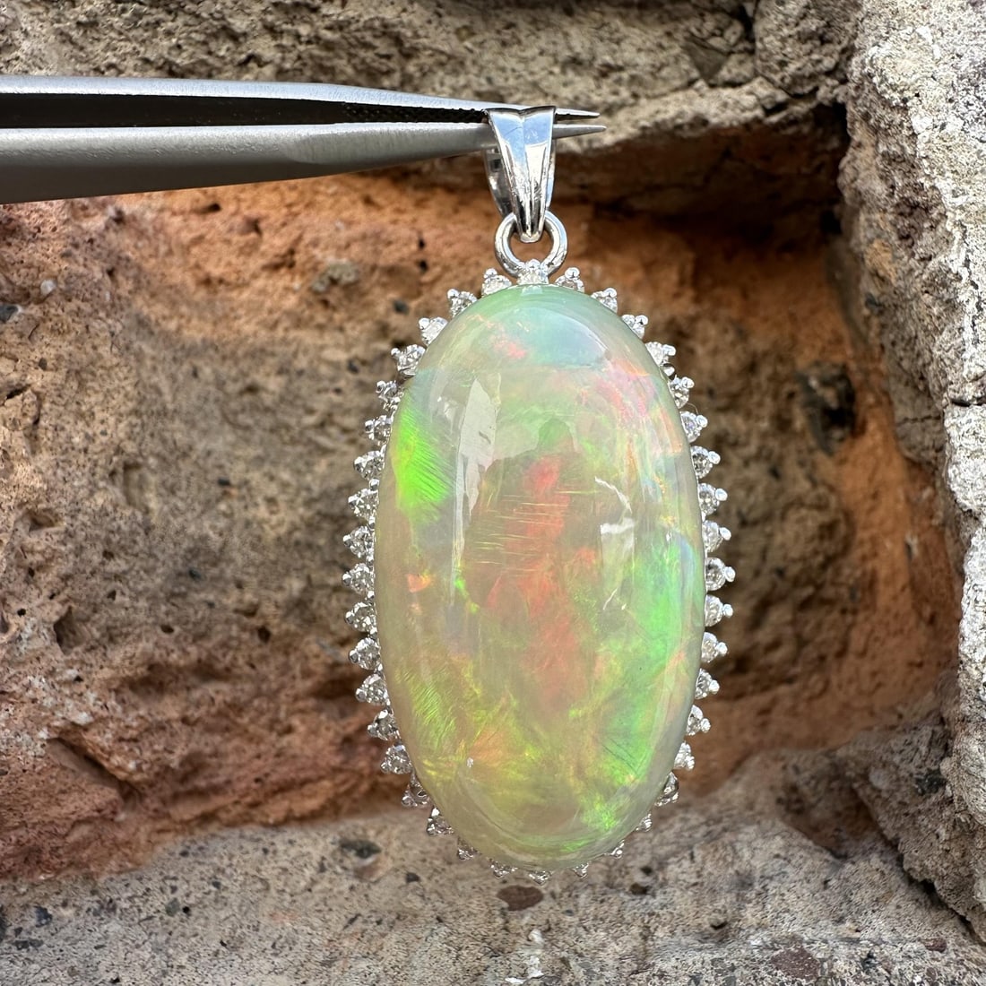 18k White Gold Opal Diamond Halo Pendant: 18k White Gold Opal Diamond Halo Pendant This elegant pendant features a large natural opal in an elongated oval cabochon cut, prong set in solid 18k white gold. The opal displays a white base color w