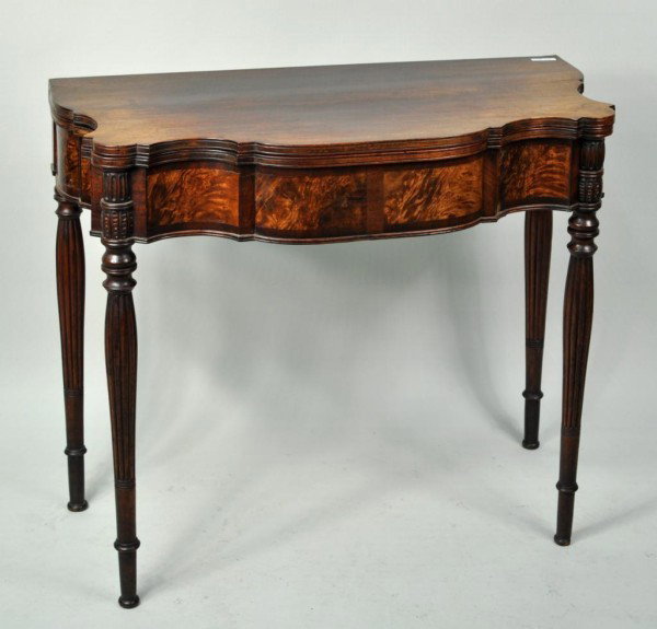 Sheraton Carved Mahogany Serpentine Card Table: Sheraton carved mahogany serpentine card table, with molded top edge above a conforming frame with brilliant figured mahogany veneers, set on turned and reeded legs with gouge carved stiles and ending