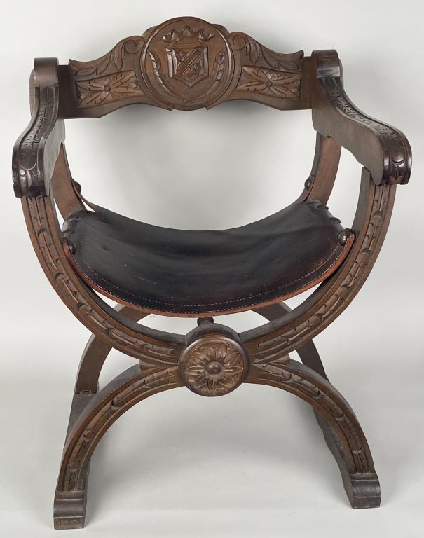 Italian Carved Walnut Savonarola Chair: with armorial carved crest and leather seat, earlier 20th century. 30" high, 24" wide, 22" deep. Wear, scuffing, and small losses to seat.