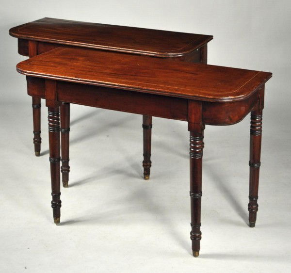 Pair of George III Mahogany Console Tables: Pair of George III satinwood inlaid figured mahogany console tables with reeded edge tops and mahogany veneered aprons, set on turned legs with brass cups. England, 19th century. Originally dining tab