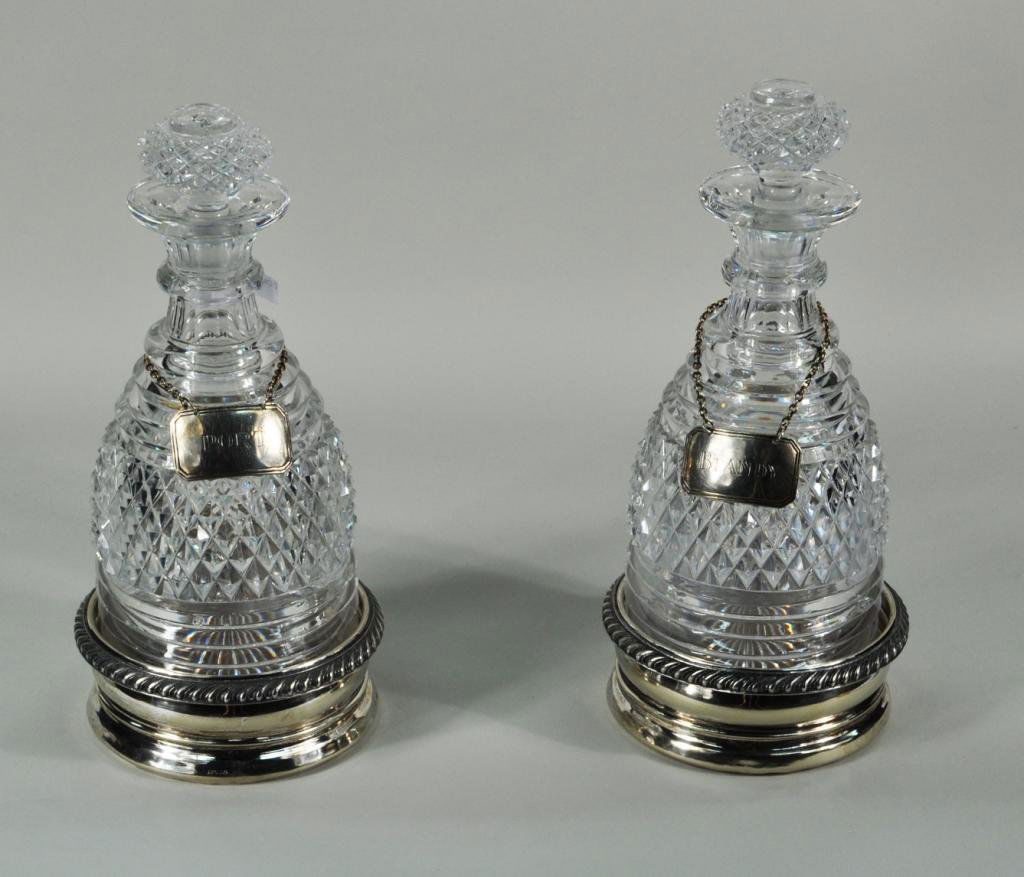 Pair Pressed Glass Decanters, Sterling Labels: Pair of pressed glass decanters with Ensko New York sterling liquor labels and two Apollo silver plate bottle coasters, chips, wear, dents to coasters, one stopper stuck. Large: 11" high, 4 1/2" diame