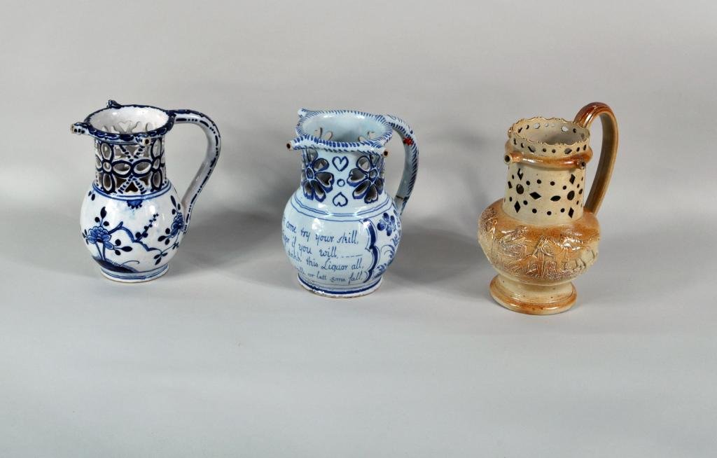Three Continental Porcelain/Pottery Puzzle Jugs: Three Continental puzzle jugs, 18th/19th century, comprising two blue and white glazed jugs and a stoneware jug with molded scenes of a hunt. Losses and repairs. Largest: 8" high, 7" wide, 5" deep; sm
