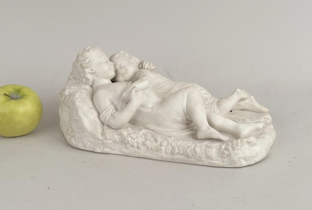 Parian Table Sculpture, Reclining Youths: oval neoclassical sculpture, two robed youths reclining in familial embrace on a leaf adorned plinth. Illegible mark on base. 5" high, 12" long, 6 1/2" deep. Soiling, apparent chip to leafage near hea