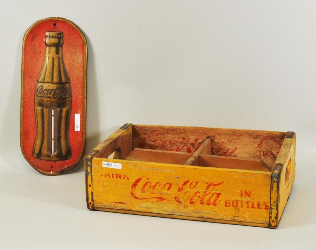 Two Coca-Cola Advertising Items: Two Coca-Cola advertising items, 20th C., comprising a lithographed metal thermometer and a stenciled bottle crate. Thermometer: 16" high, 6 3/4" wide, crate: 12" high, 18" wide. Condition: both with 