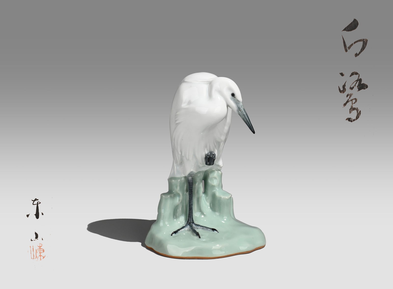 White Heron Ceramic Okimono by Miyanaga Tozan: This porcelain figure depicts a white heron standing gracefully upon a stylized rock, rendered with refined naturalism and poetic elegance. The bird’s slender body curves gently as it lowers its hea