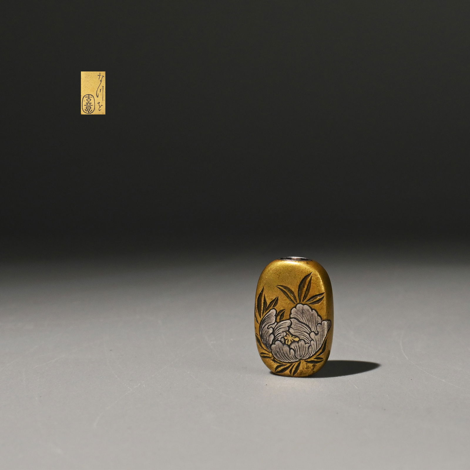 Copper ojime with silver inlay, by Kano Natsuo: This is a copper ojime by Kan? Natsuo, decorated with a peony motif in silver inlay. The warm-toned copper ground is contrasted with finely inlaid silver, while the peony’s petals and leaves are