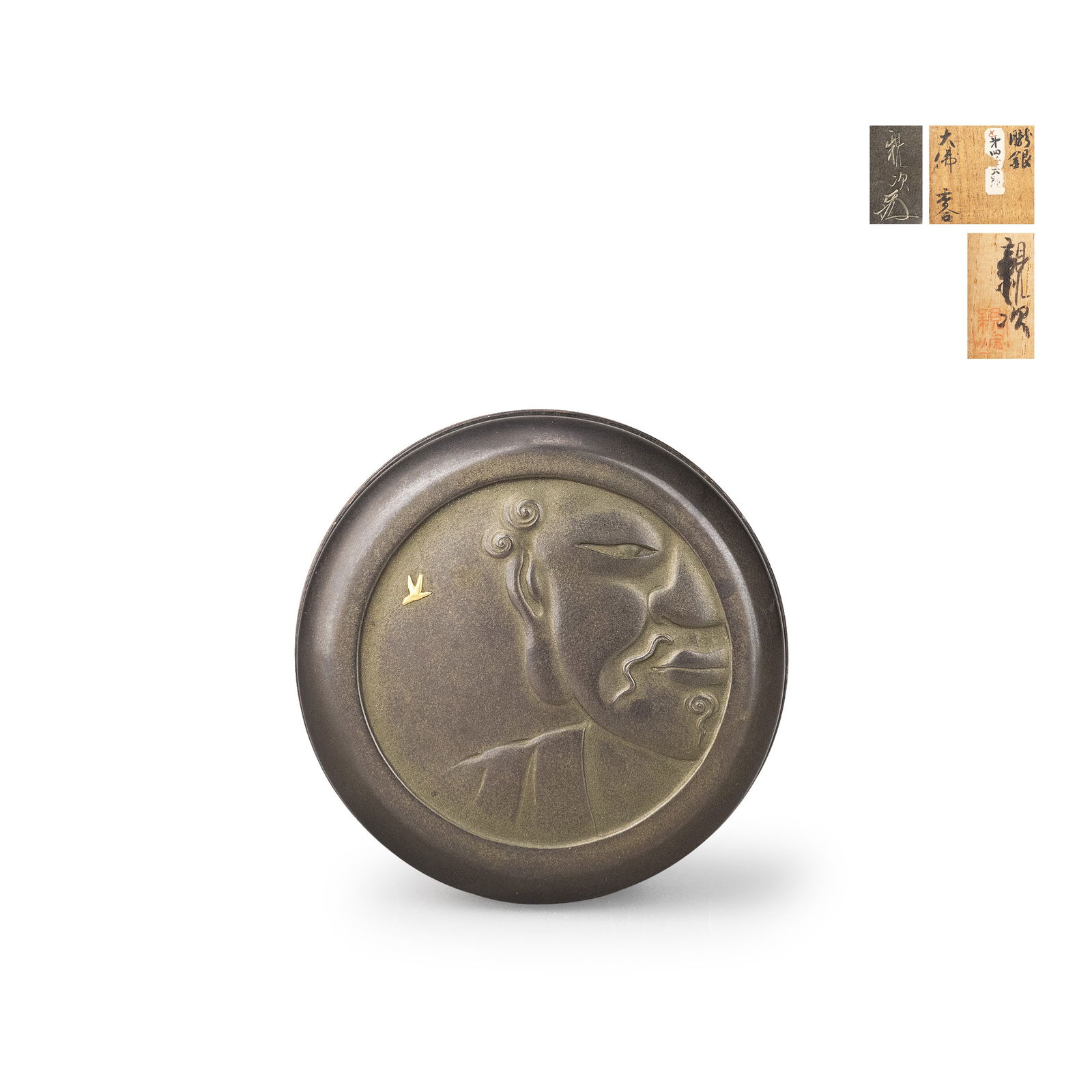 Incense Container with Gold Inlay by Shinji Shiozu: A circular incense box crafted in shibuichi, the subdued grey-brown surface serving as ground for a high relief depiction of the serene face of the Great Buddha. The features are rendered with restrai