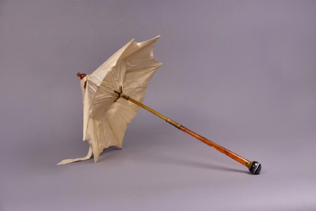 SILK PARASOL with AMBER and AGATE HANDLE, LATE 19th C.: Ivory silk faille canopy with folding amber carriage handle and bone stick with brass collar, terminating in a black and white agate ball decorated with brass filigree. Overall L-24, canopy-16. (Canop