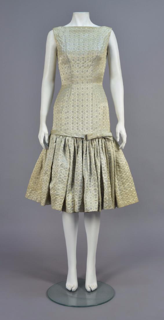SUZY PERETTE METALLIC BROCADE COCKTAIL DRESS, 1950s: Tone-on-tone platinum brocade with repeating roundels and tiny metallic gold crosses, sleeveless boatneck fitted through the hip, full lower skirt having self band with bow. Labeled. B-34, W-22, H-34,