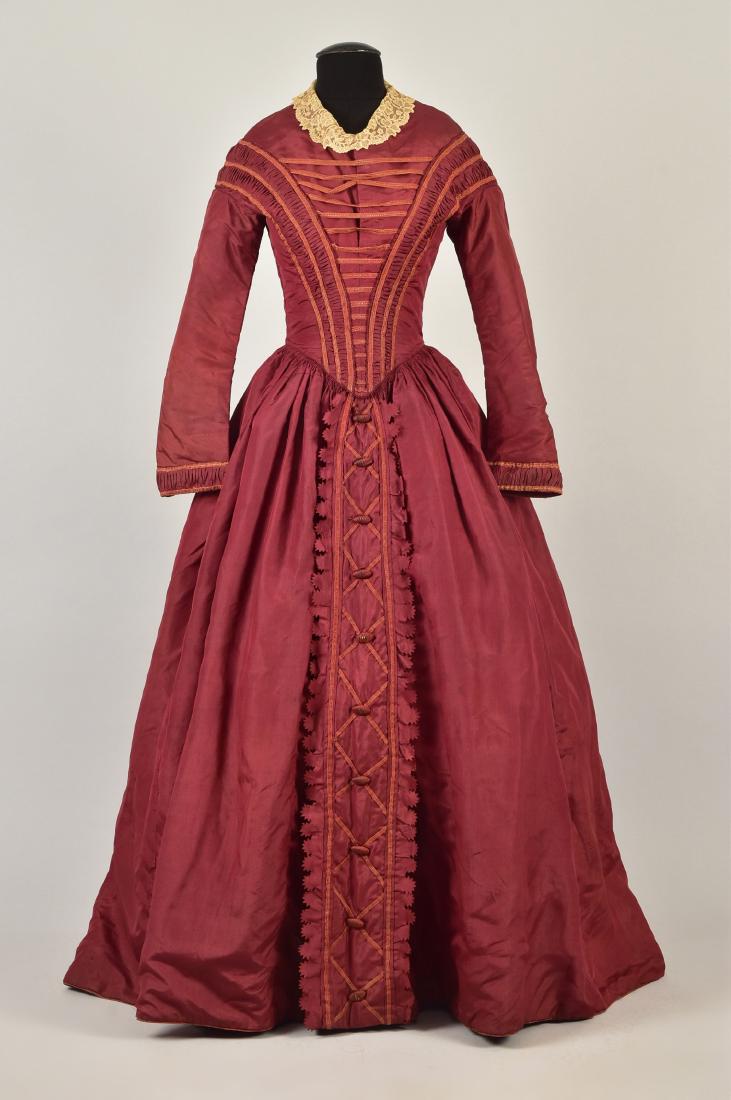 ENGLISH CLARET SILK DAY DRESS, 1845: Long sleeve bodice trimmed with pleats and silk braid, having front point and cream lace collar, full skirt with gauged gathers, the center panel decorated with braid and ribbed oval domes flanked by 