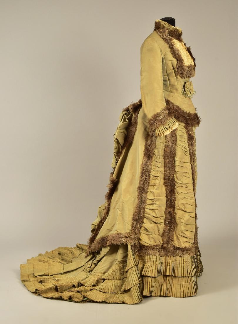 OSTRICH FEATHER-TRIMMED BUSTLE GOWN, 1876: 2-piece grey-faune taffeta faille, having long sleeve peplum bodice with pleated collar and cuff, decorated with self bows, cream bobbin lace and feathers; trained skirt with vertical ruched panels to