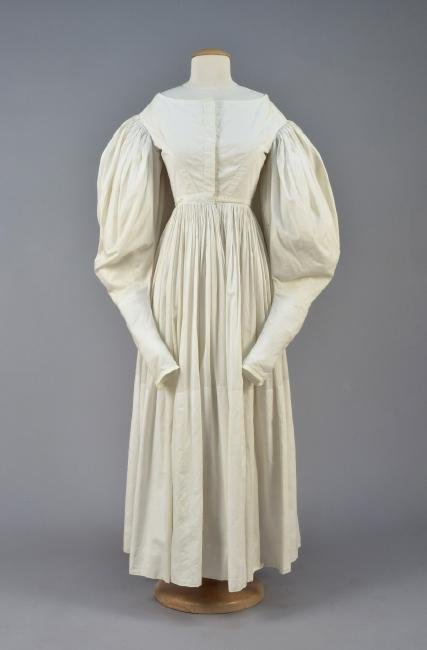 COTTON GOWN with IMBECILE SLEEVES, 1830s: White boatneck having long sleeve tightly-gathered into self piping at the armhole, curved bodice seams and hidden patterned glass buttons, waistband with tie, full gathered skirt with very deep hem. 