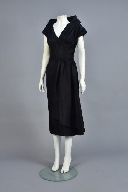 JACQUES FATH ASYMMETRICAL COCKTAIL DRESS, 1952: Black silk taffeta with small cap sleeve, asymmetrical crossed front panels with back stand-up collar, the left side gathered behind the front and joined with faux self buttons, side closure with hook