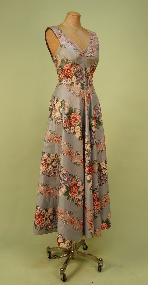 FLORAL PRINTED CHINTZ SUMMER GOWN, 1957. Sleeveles: FLORAL PRINTED CHINTZ SUMMER GOWN, 1957. Sleeveless V-neck with wide straps having red, pink, plum, green and white floral bands on pale grey, nine self button and loop back closure, bias cut near ful